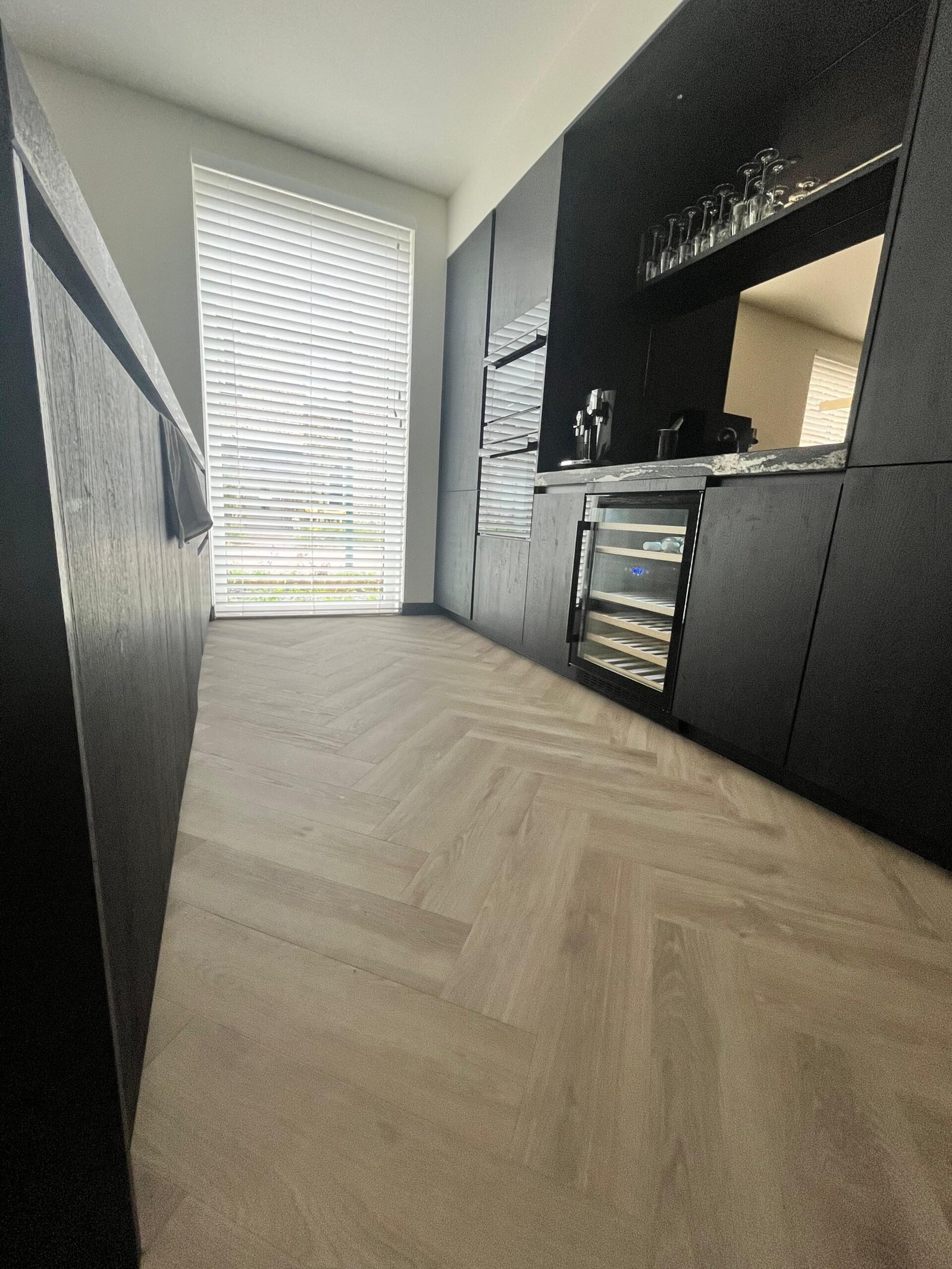 Een grijs beige visgraat vloer in een ruime zwarte keuken met een groot raam.